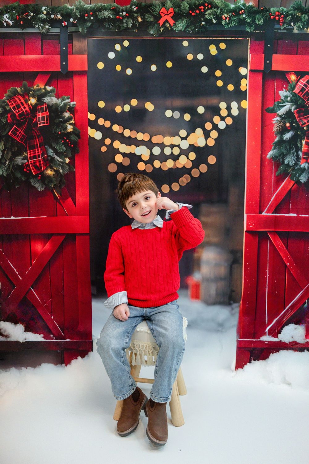 Festive Christmas scene with red barn doors, wreaths, string lights and snow, featuring a bright red sweater against wooden backdrop.