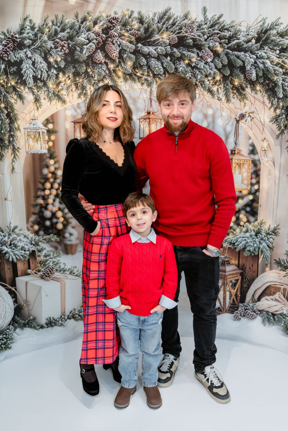 Family wearing coordinated red and black holiday outfits poses in front of snowy winter backdrop with frosted branches.