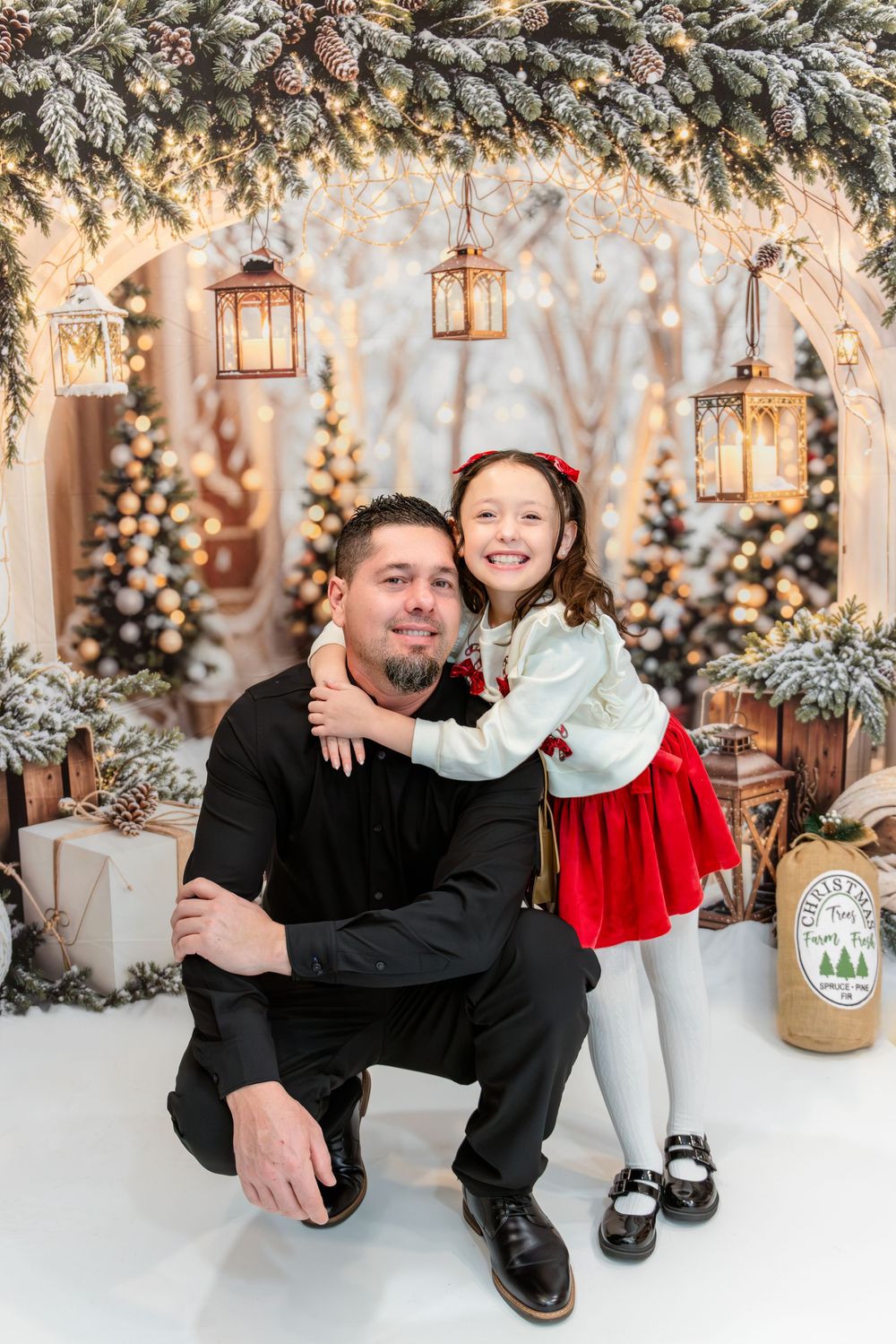 Festive winter photo session with snowy Christmas decor, lanterns and decorated evergreen trees in the background.