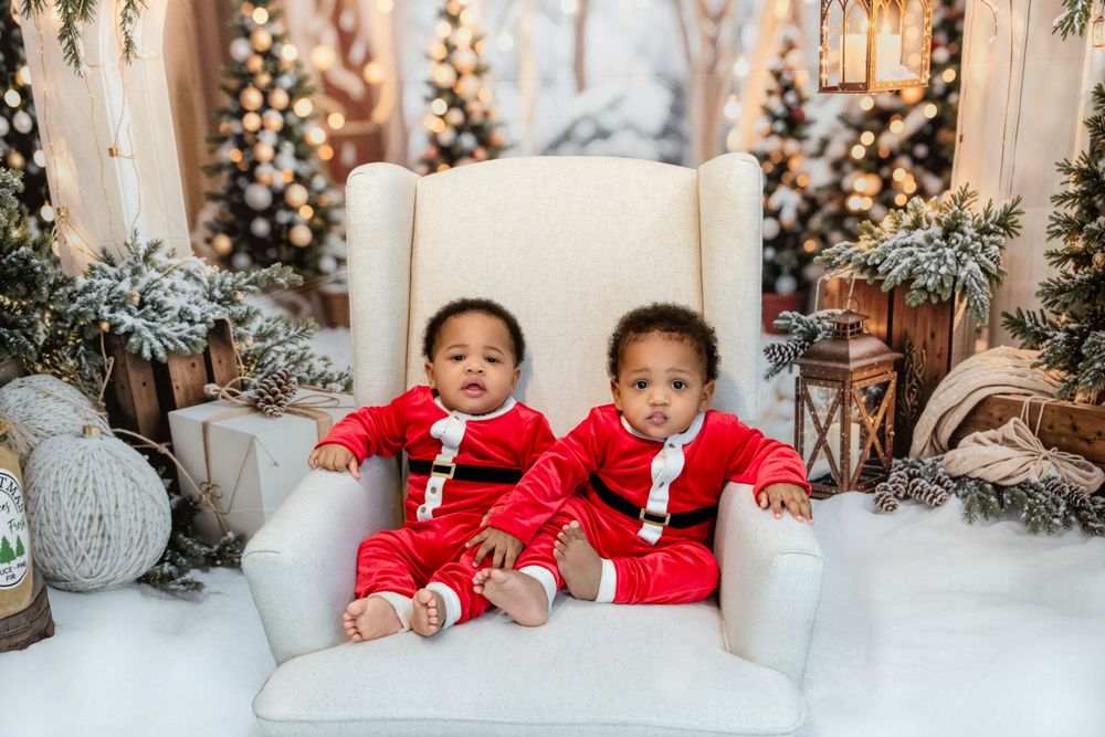 Two children in matching red Santa outfits sit on a white chair surrounded by decorated Christmas trees and winter decor.