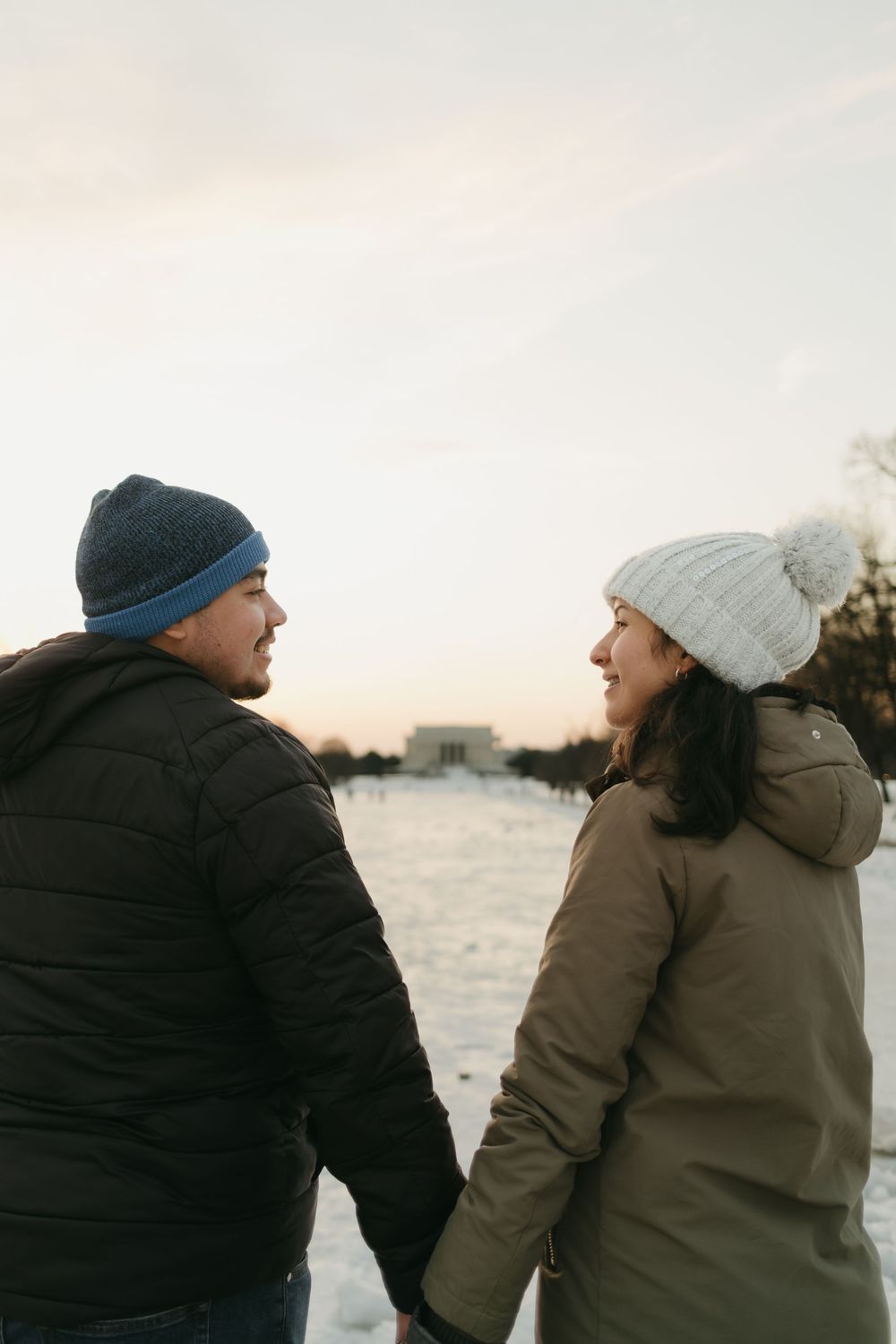 A couple in winter coats and beanies sharing an intimate moment against a snowy landscape at sunset.
