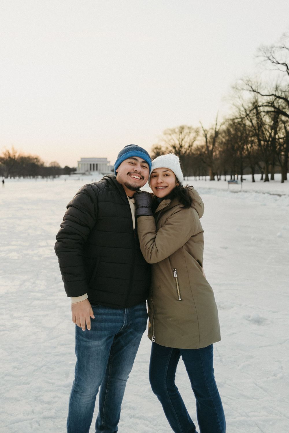 A couple in winter coats and knit hats embrace while standing in a snowy park at sunset.