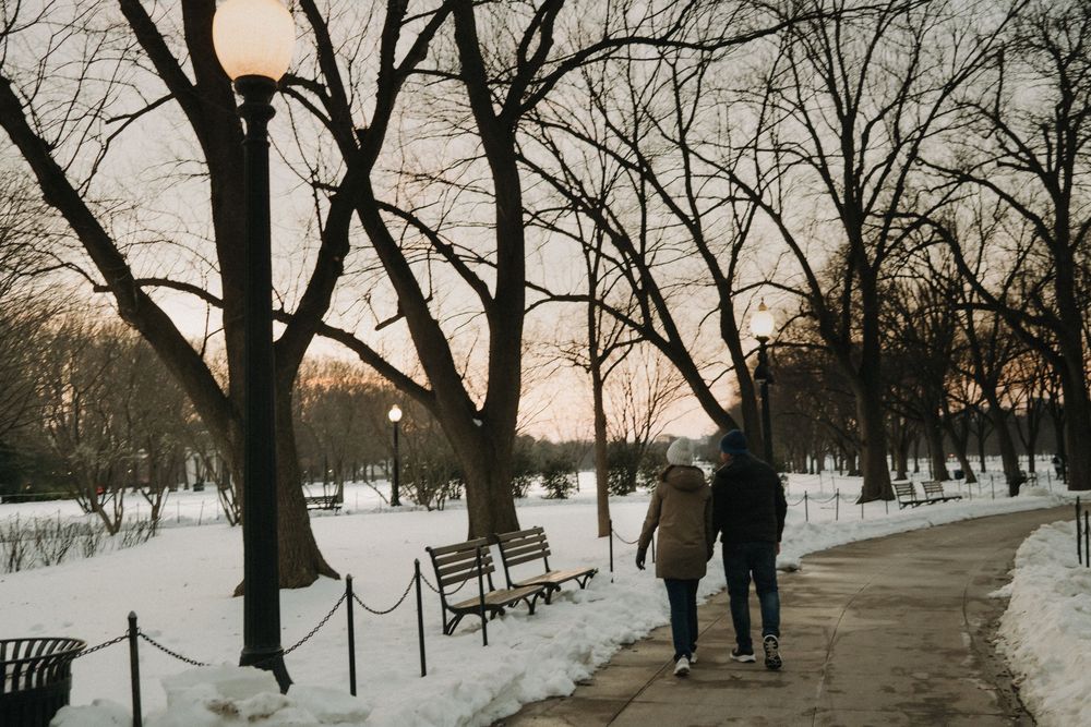 A couple walks along a snow-lined path at sunset with bare winter trees silhouetted against the sky.