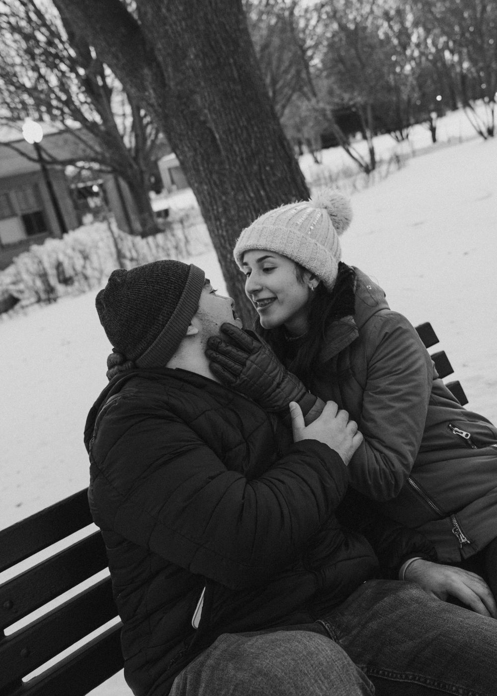 Two people sitting close together on a park bench in winter, sharing a playful moment in the snow.