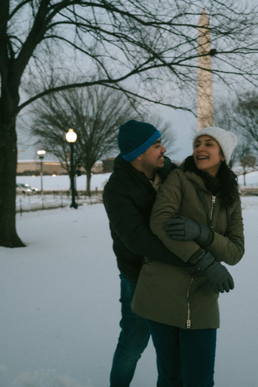 A couple in winter coats embraces and shares a playful moment in a snowy park landscape.
