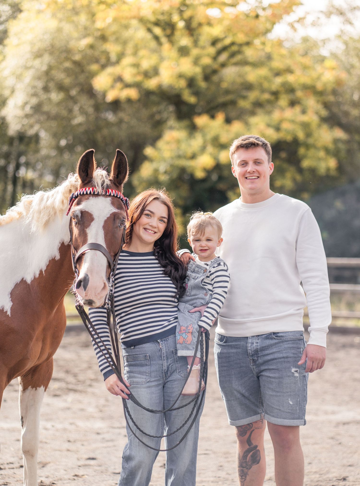 A family in matching striped shirts and denim poses with their paint horse in a sunny outdoor setting.