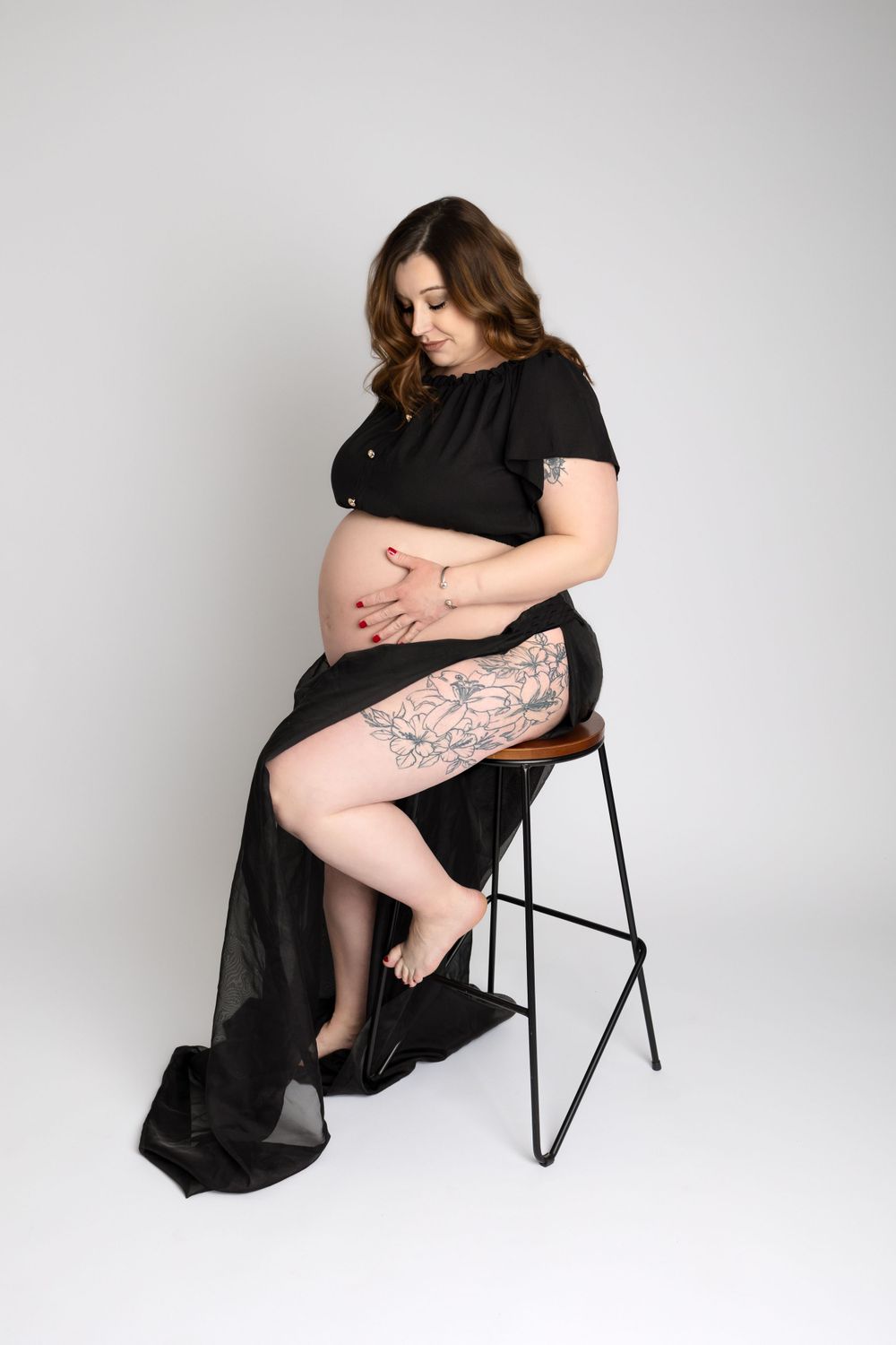 Artistic maternity photo of woman in black dress seated on stool showing tattoo.