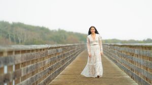 Person in white dress standing centered on long wooden boardwalk with trees in background.