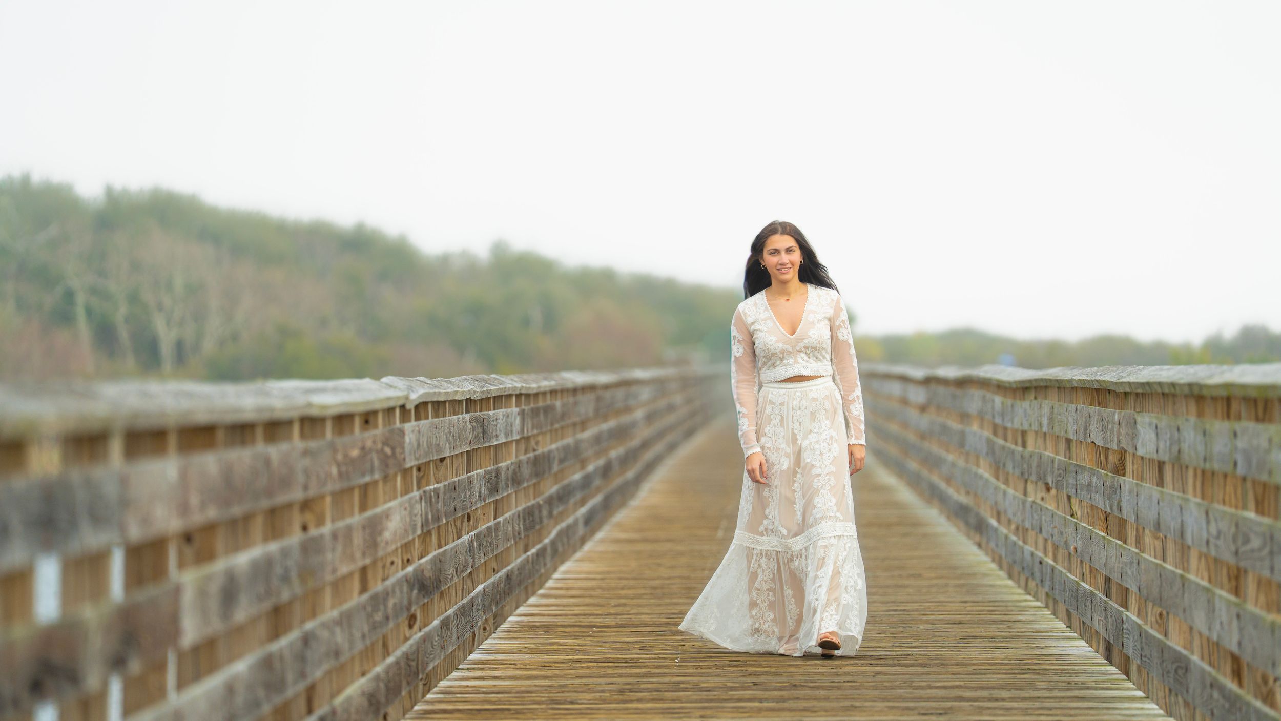 Person in white dress standing centered on long wooden boardwalk with trees in background.