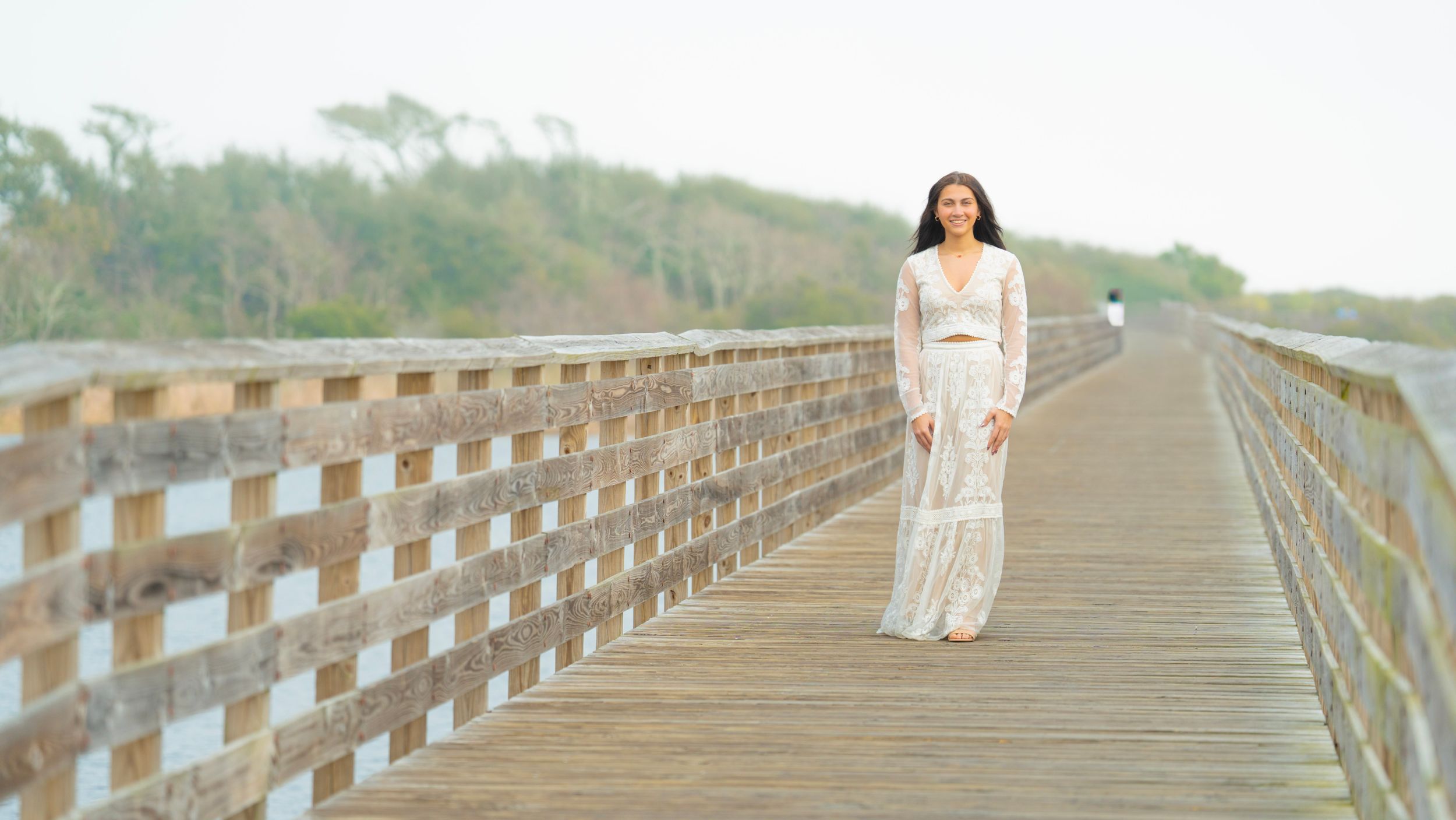 Figure in white flowing dress walking on wooden boardwalk surrounded by nature.