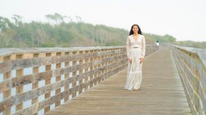 Figure in white flowing dress walking on wooden boardwalk surrounded by nature.