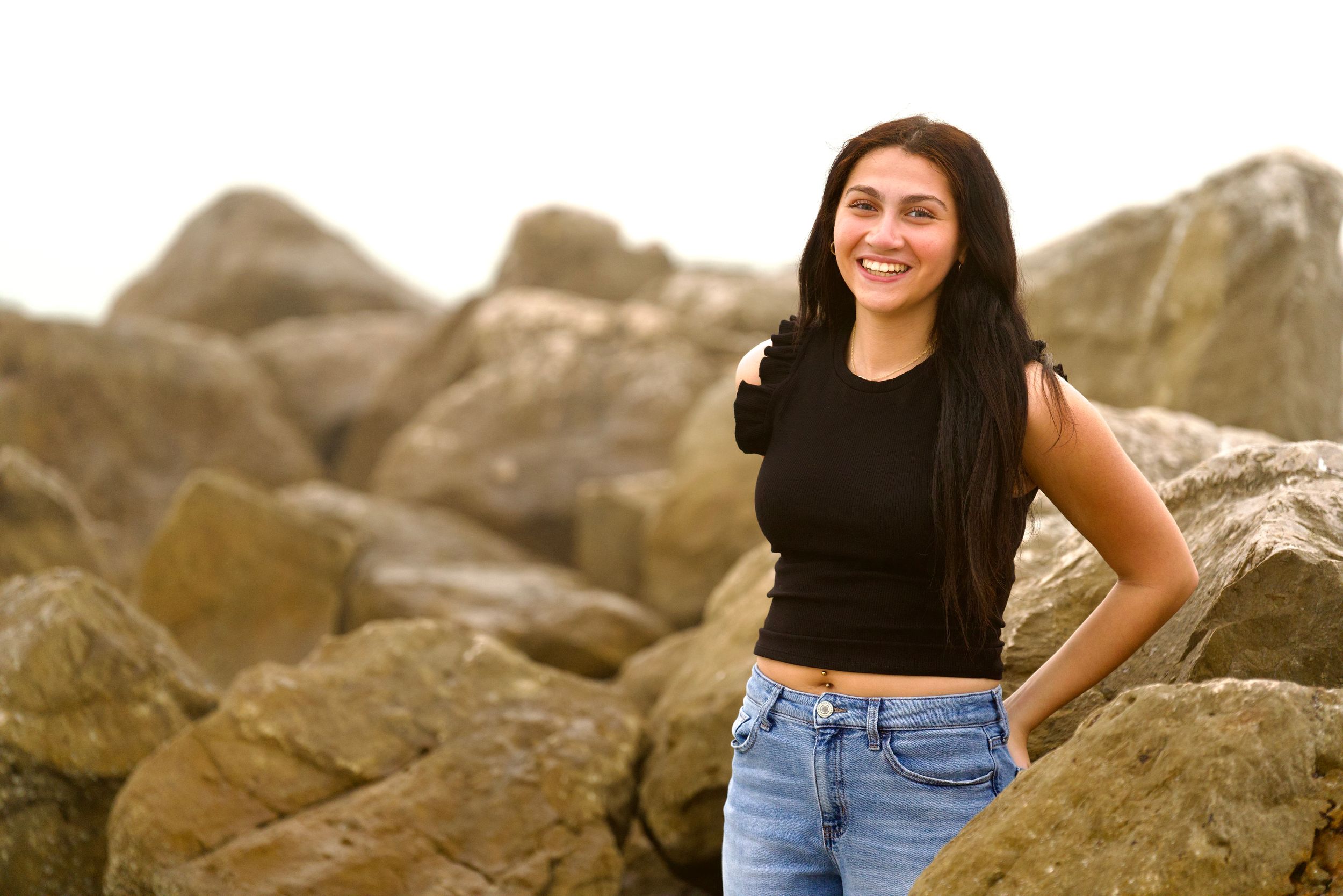 Person in black crop top and jeans posing confidently on rocky beach formation.