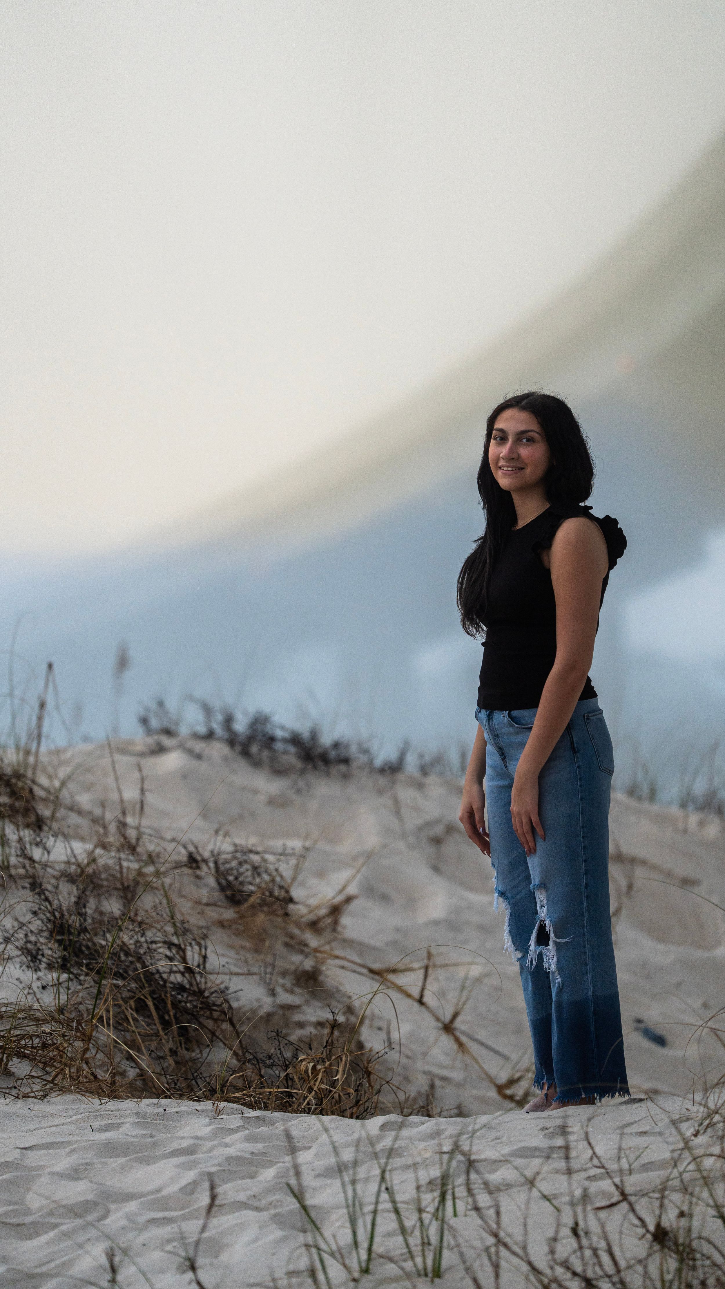 Figure standing on sandy dunes wearing black top and jeans against cloudy sky.