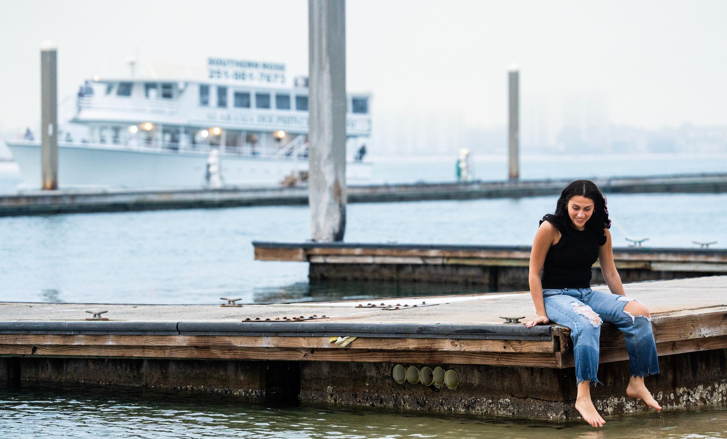 Person sitting on wooden dock near water with boat in background.