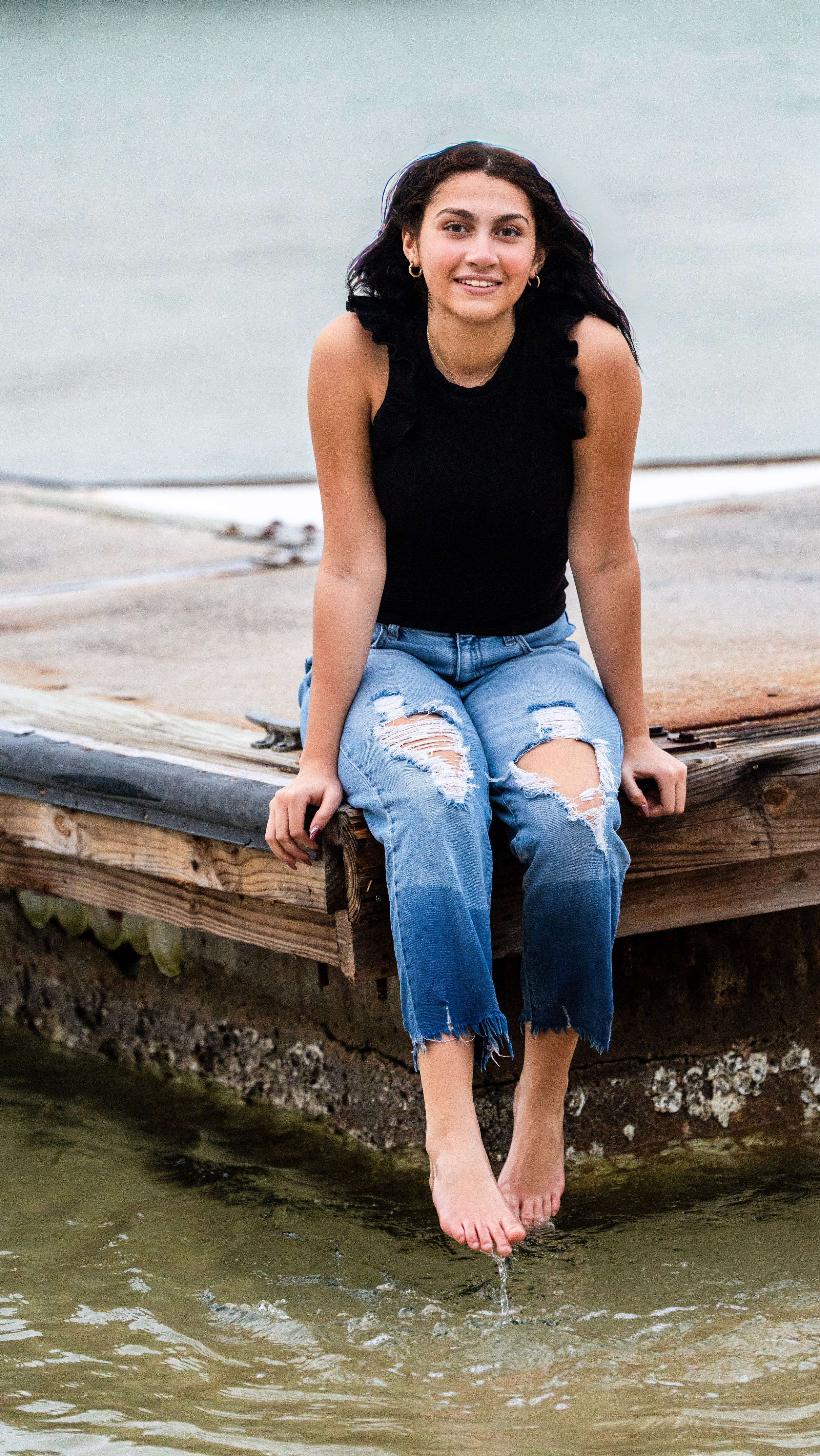Person in black top and ripped jeans sitting on wooden dock edge near water.