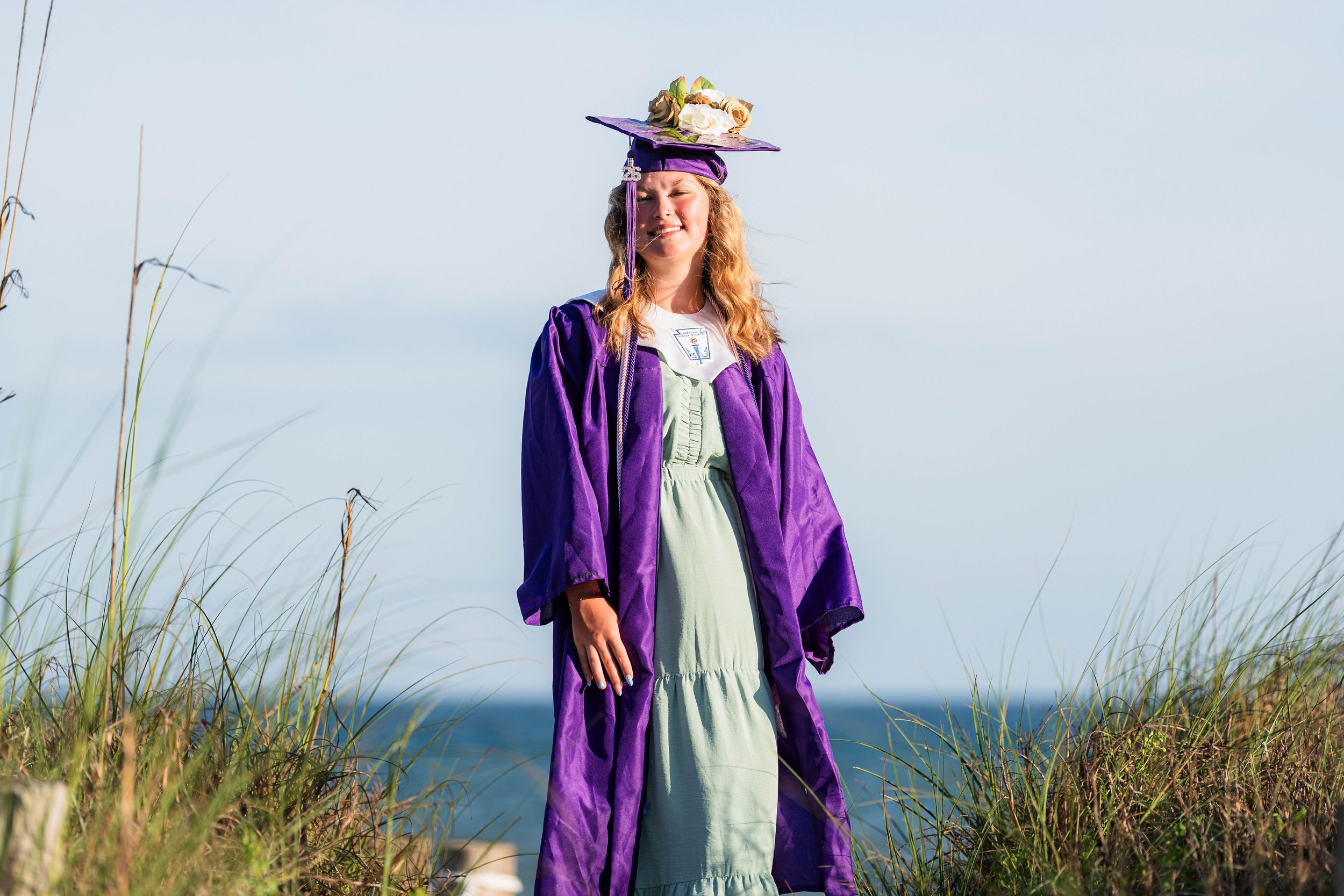 Young woman in purple graduation gown and cap with flowers stands on beach dunes with ocean in background.