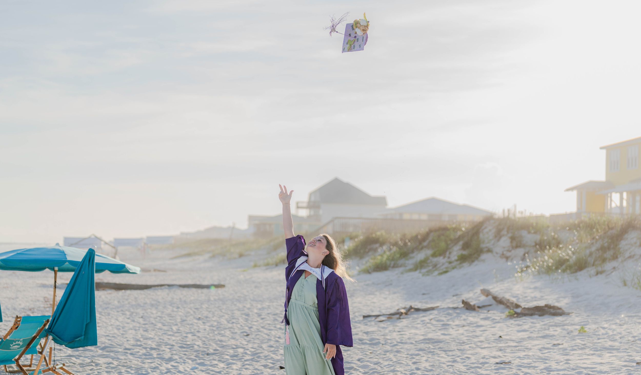 Graduate in cap and gown tossing graduation cap on a sunny beach with ocean backdrop.