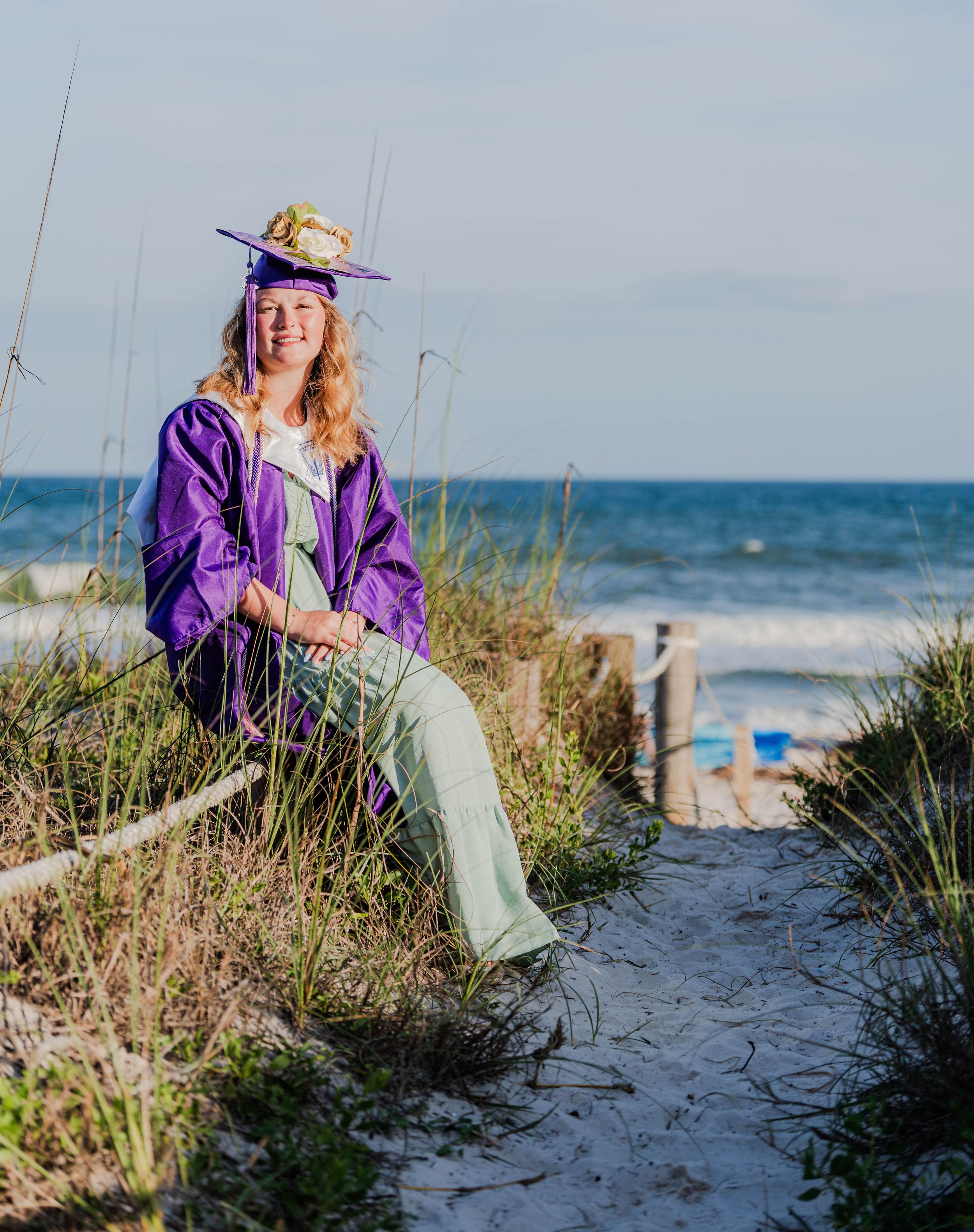 A smiling graduate in purple cap and gown sits on a sandy beach path surrounded by sea oats on a sunny day.