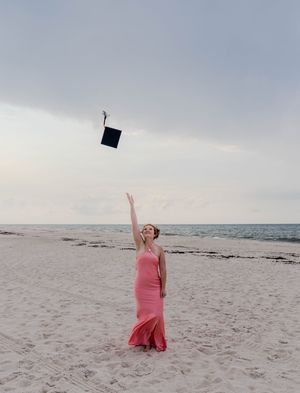 A woman in a pink gown tosses her graduation cap into the air on a sandy beach at dusk.