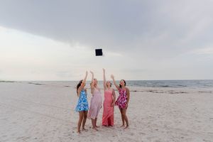 Four women in colorful dresses celebrate on a beach, tossing a graduation cap into the air.