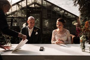 Wedding couple signs marriage documents at an outdoor ceremony with sunlight streaming through trees.