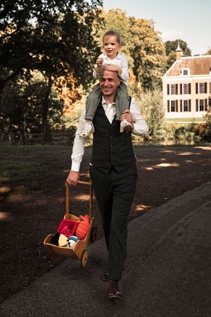 A person in formal attire walks on a path pulling a red wagon while carrying a child on their shoulders.