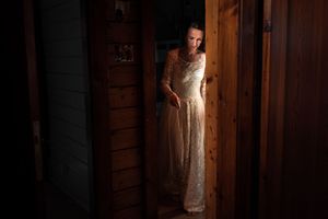 A bride in a flowing white lace wedding dress poses in a rustic wooden barn setting with dramatic lighting.