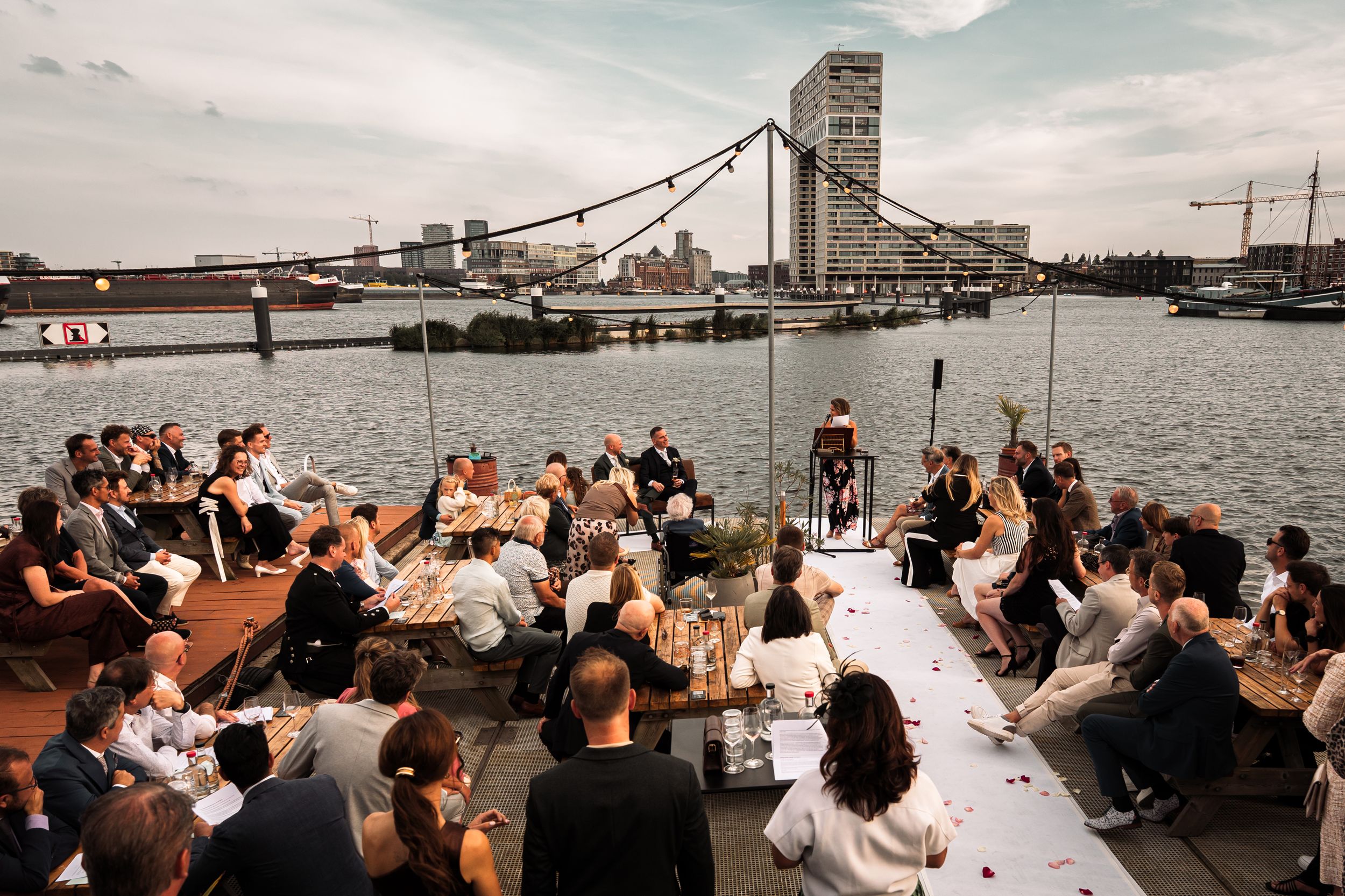 A panoramic view of a waterfront wedding reception with guests seated at long tables overlooking a harbor.