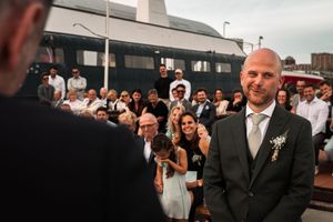 A group of wedding guests gathered outdoors watching something off-camera with excited expressions.
