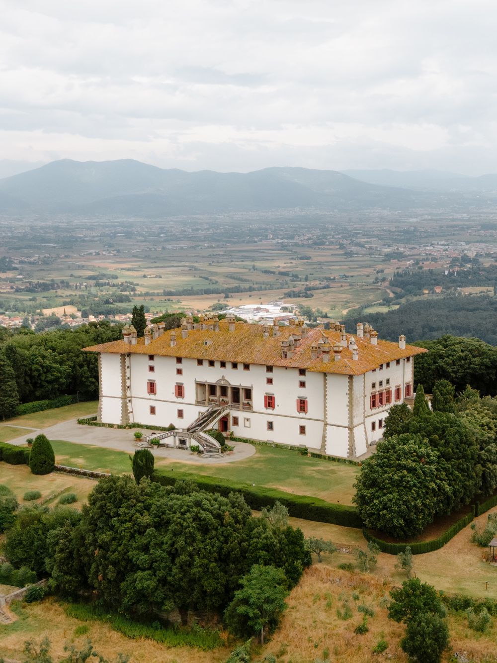 Aerial panoramic view of a historic white monastery complex nestled on a hilltop overlooking scenic countryside. Tenuta di Artimino