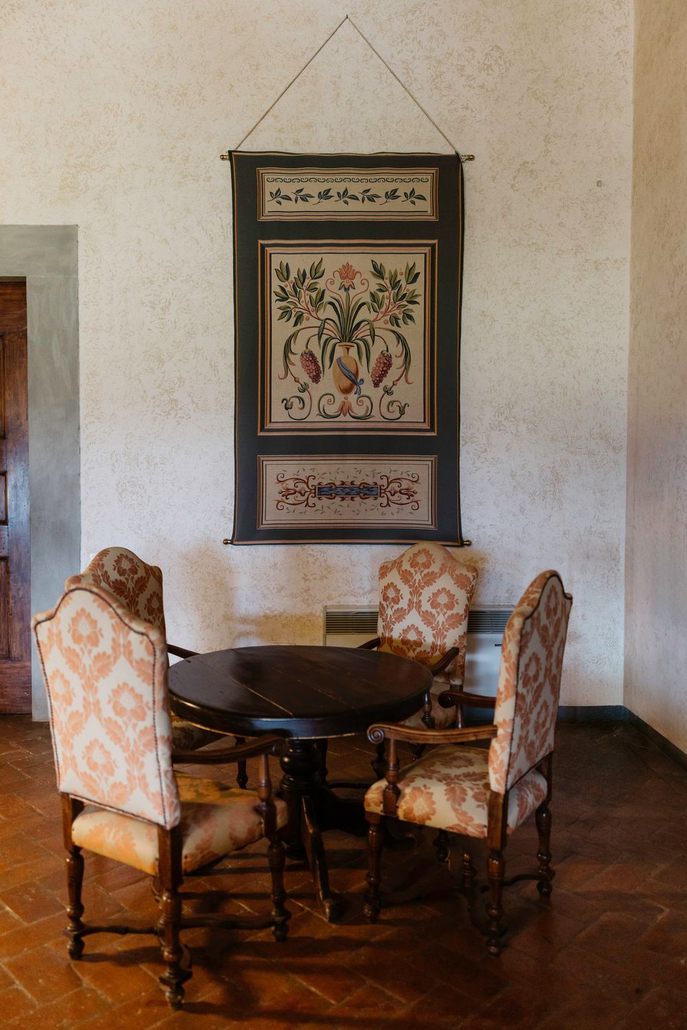 Antique wooden chairs with floral upholstery arranged around a dark table beneath decorative wall hangings in a rustic room.