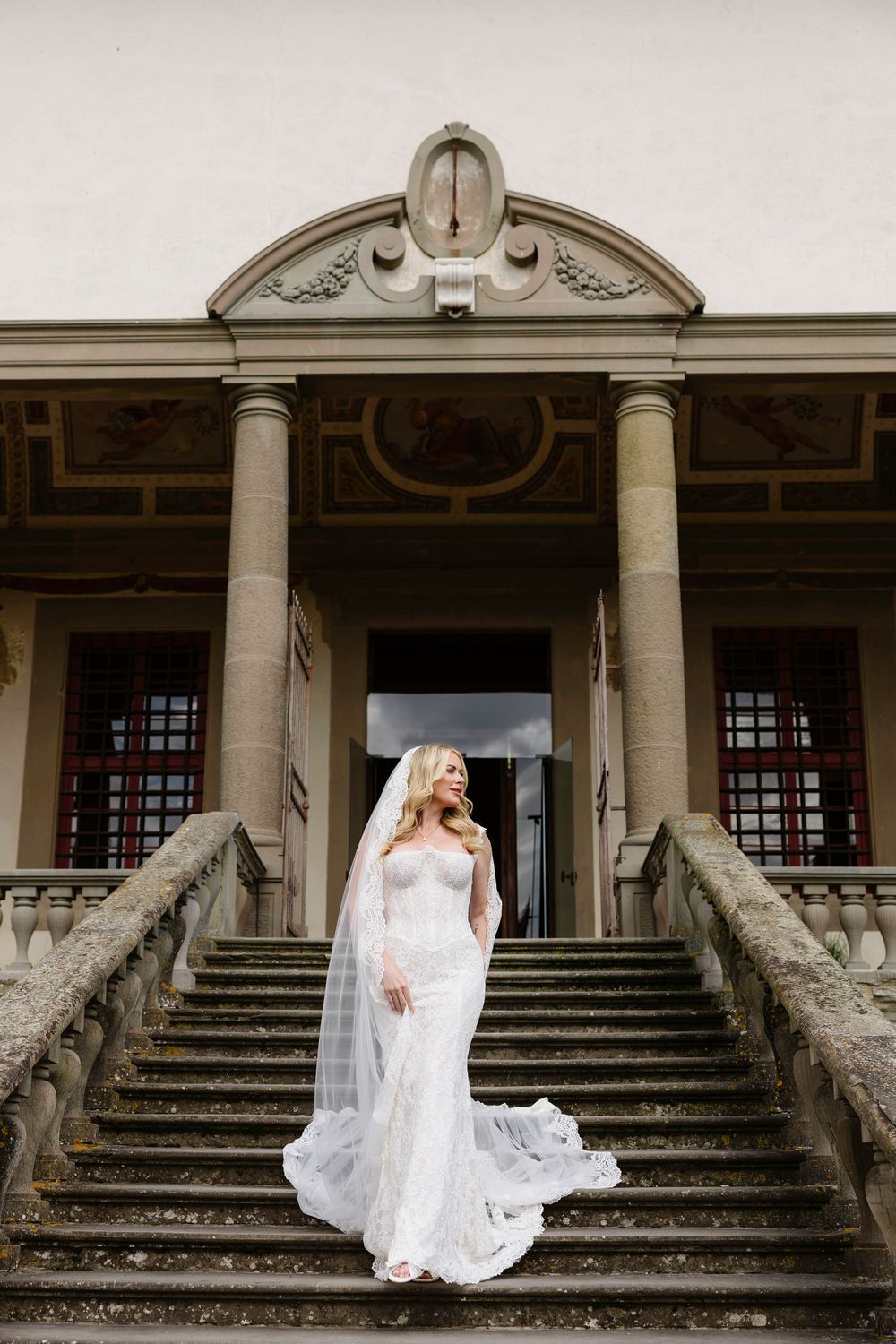 Elegant bride in white lace gown and flowing veil stands on grand stone staircas of historical villa Tenuta di Artimino Tuscany