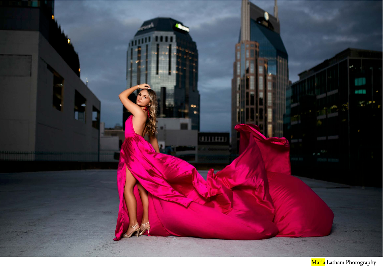 A flowing hot pink evening gown billows dramatically against a moody urban rooftop setting with skyscrapers in the background.