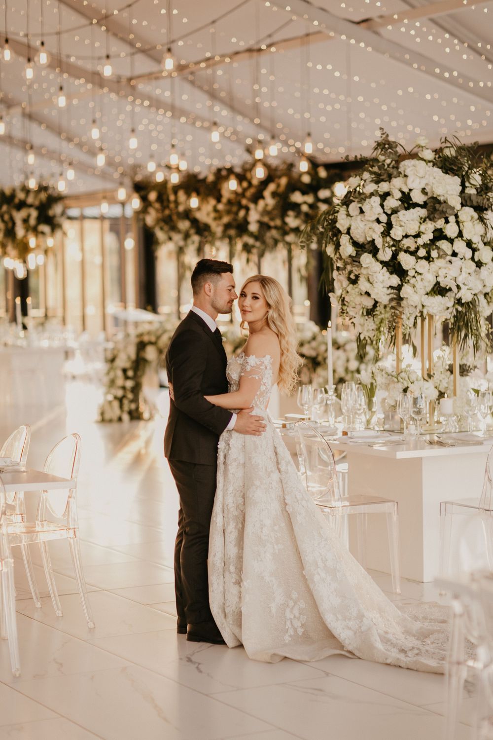 Bride and groom embracing in elegant white floral wedding reception venue with string lights.