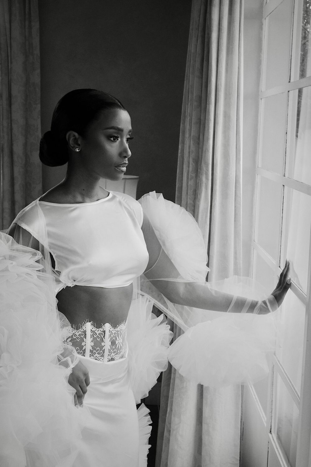 Black bride in white crop top and tulle skirt gazing out window in black and white portrait.