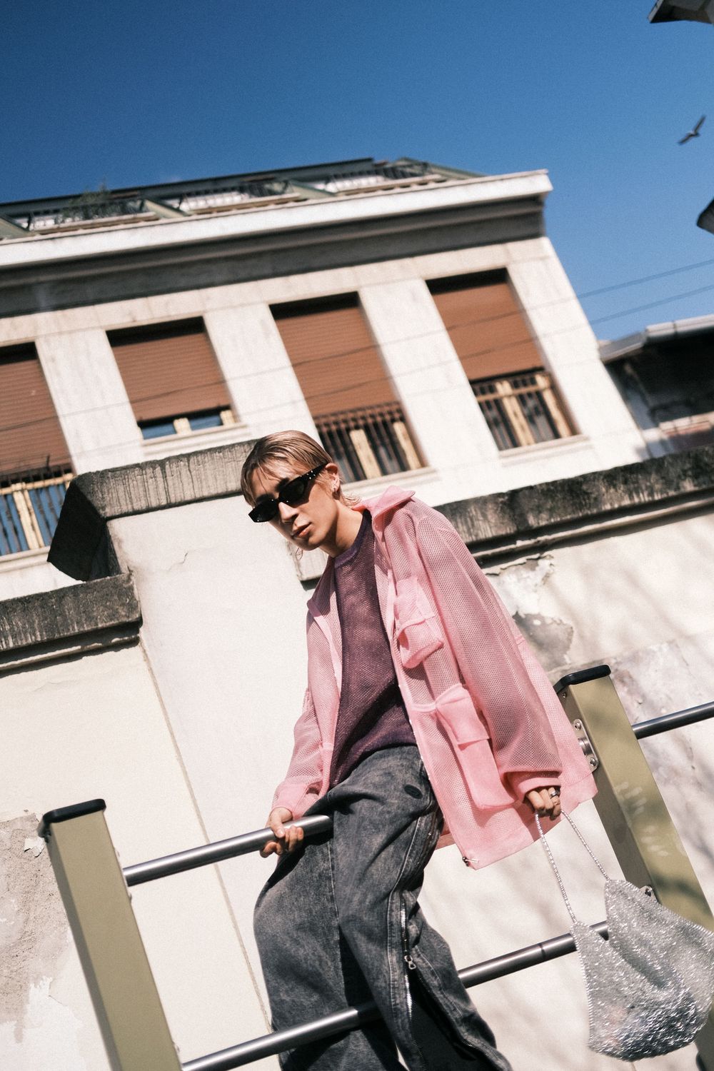 Person in pink blazer and sunglasses poses against white wall in urban setting with angled architectural elements.