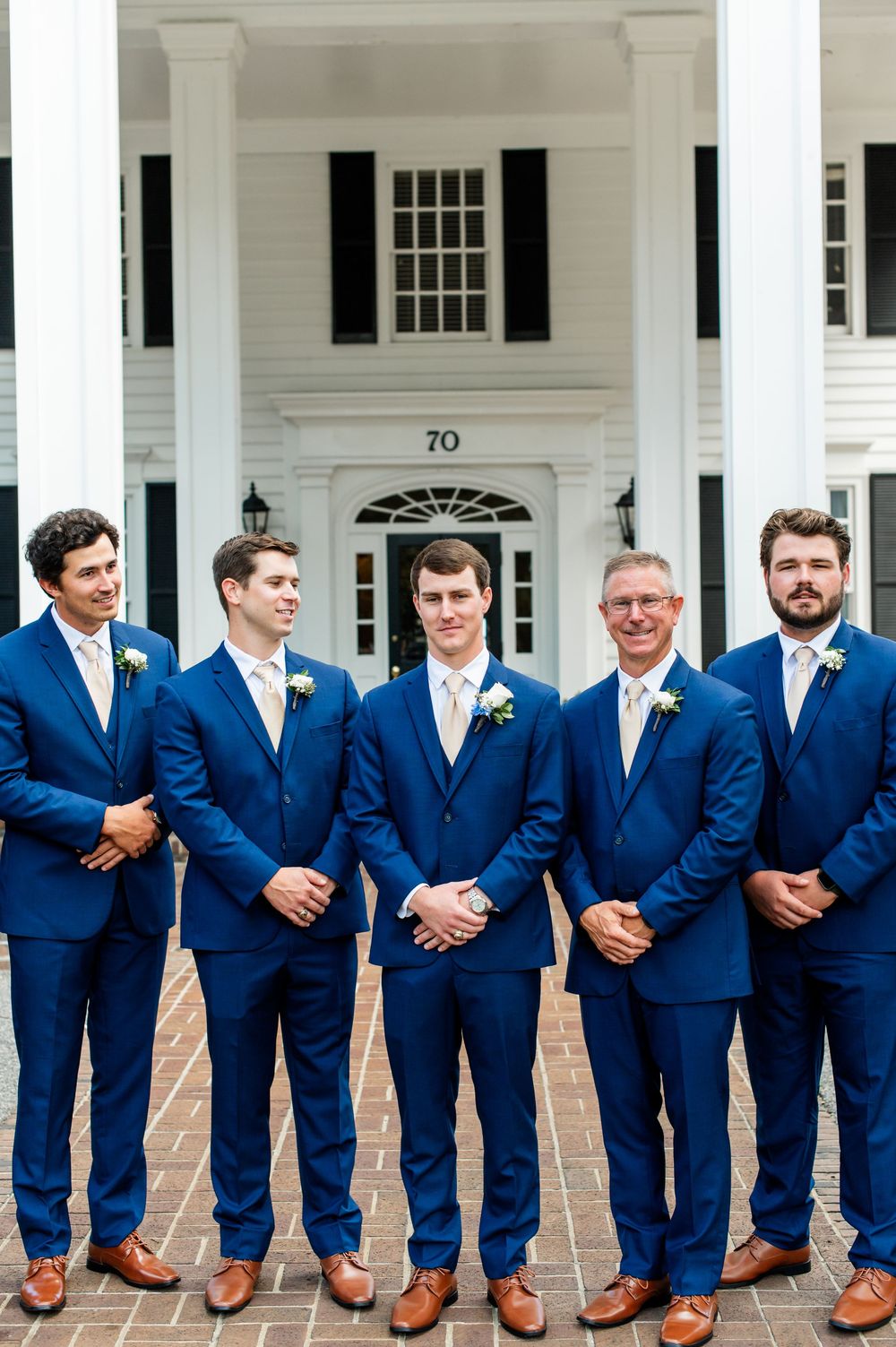 Groomsmen in matching navy suits and brown shoes line up against white colonial building.