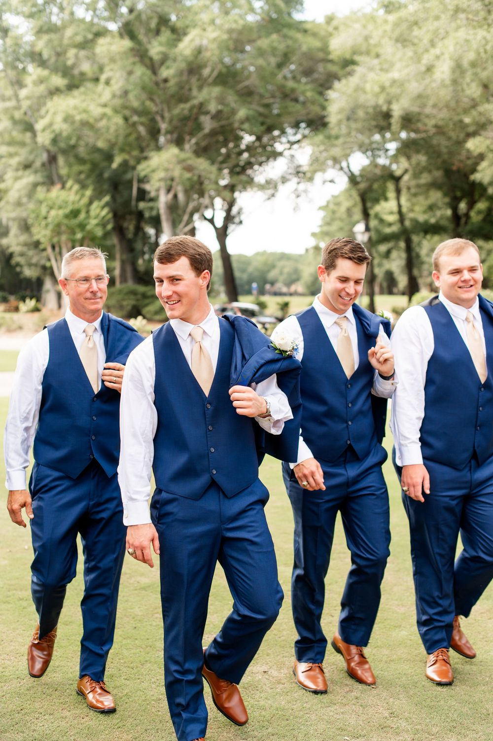 Fun groomsmen photo showing synchronized walking poses in navy suits on grass.