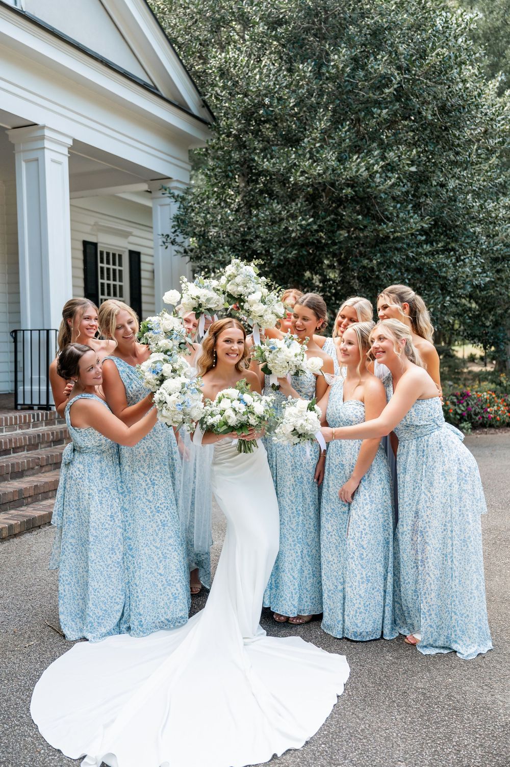 Wedding party in sparkly blue dresses and white gown gather for candid photos outside a white building.