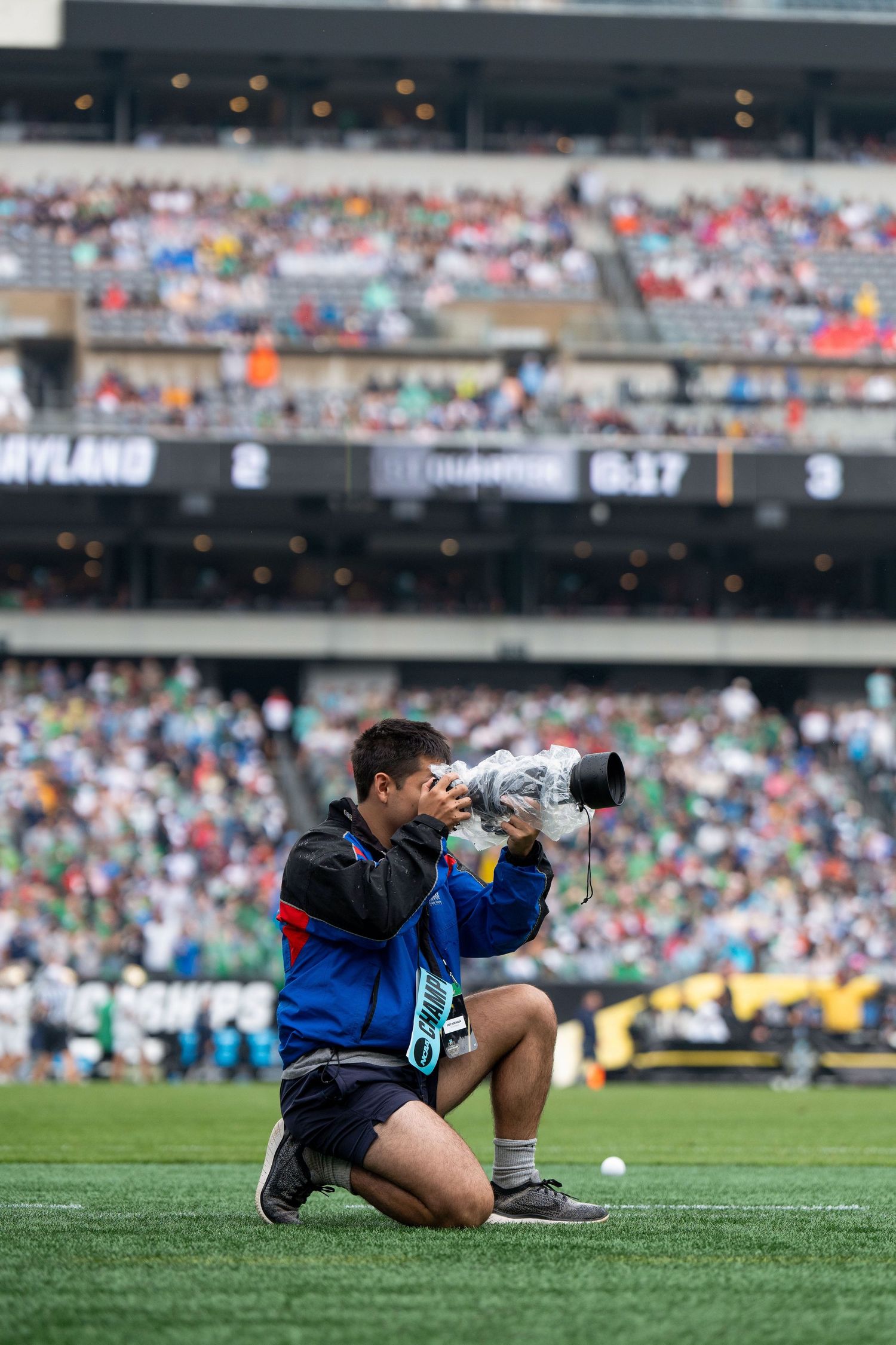 Sports photographer lying on grass field capturing action shots during game at crowded stadium.