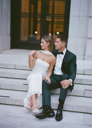 Couple in wedding attire sitting casually on stone steps of a grand building.