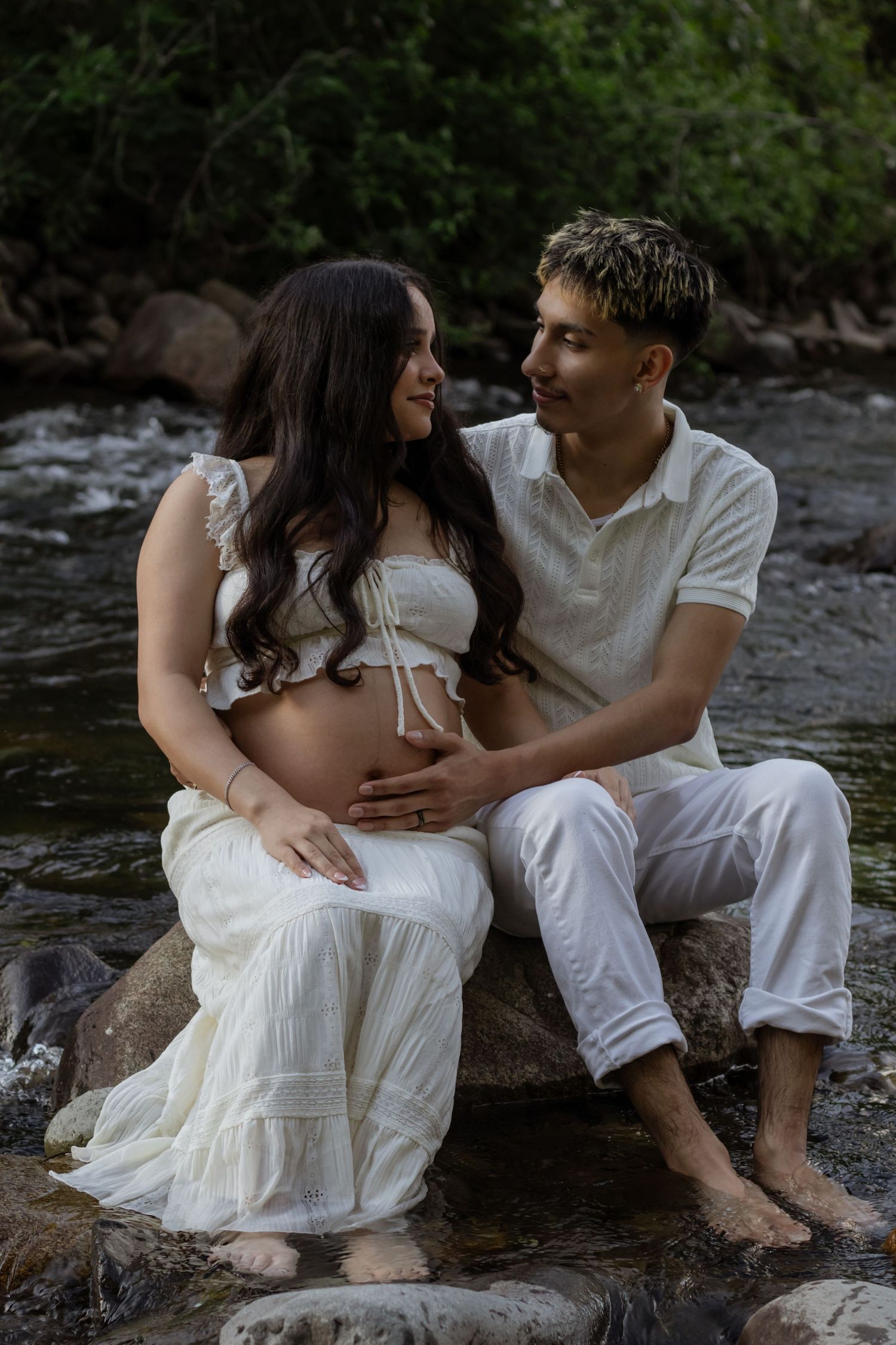 Pregnant couple in white outfits sitting on rocks by little mashel river, sharing a tender moment while cradling baby bump.