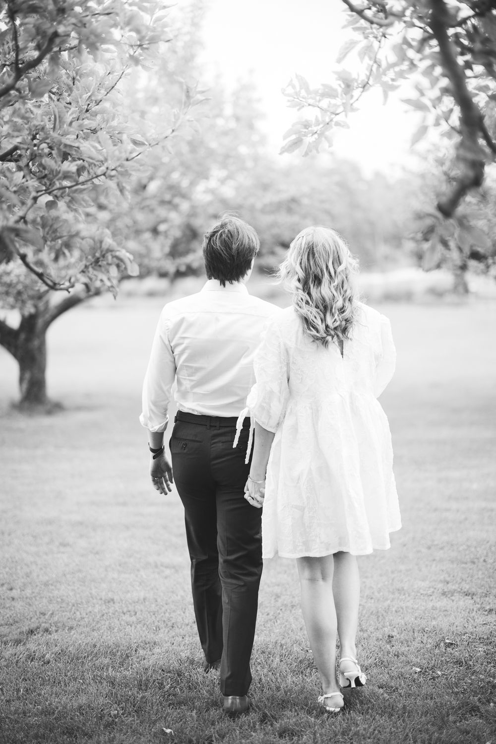 Black and white photo of a couple walking hand in hand through an orchard in soft focus.