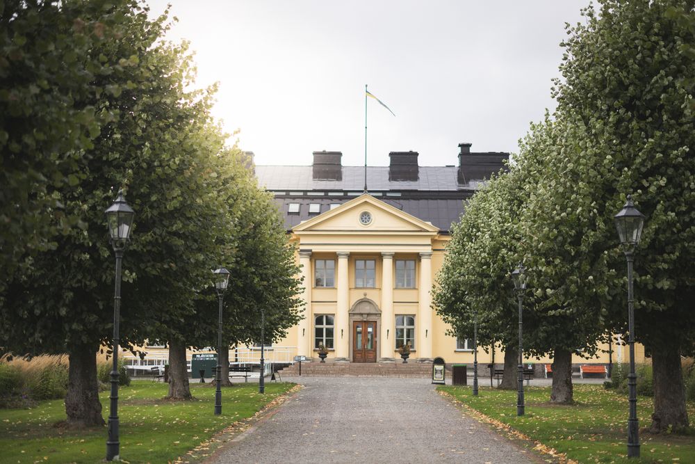 A panoramic view of a historic yellow building with trees lining the path leading to its entrance.