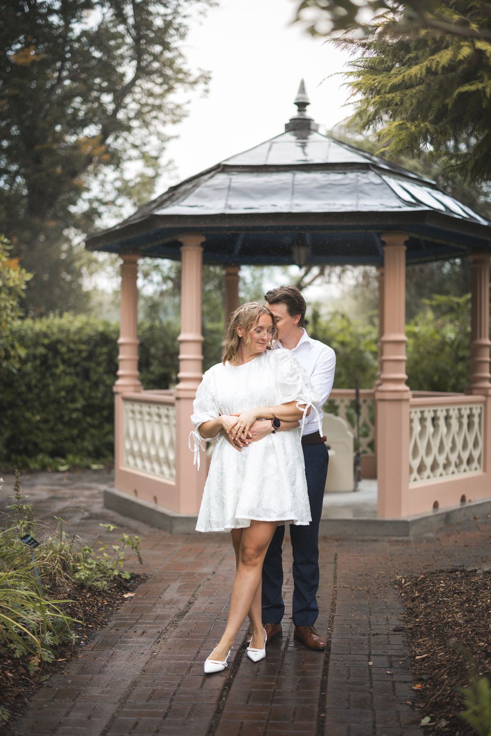 A romantic couple poses together in a garden gazebo setting with brick pathways and ornate pillars.