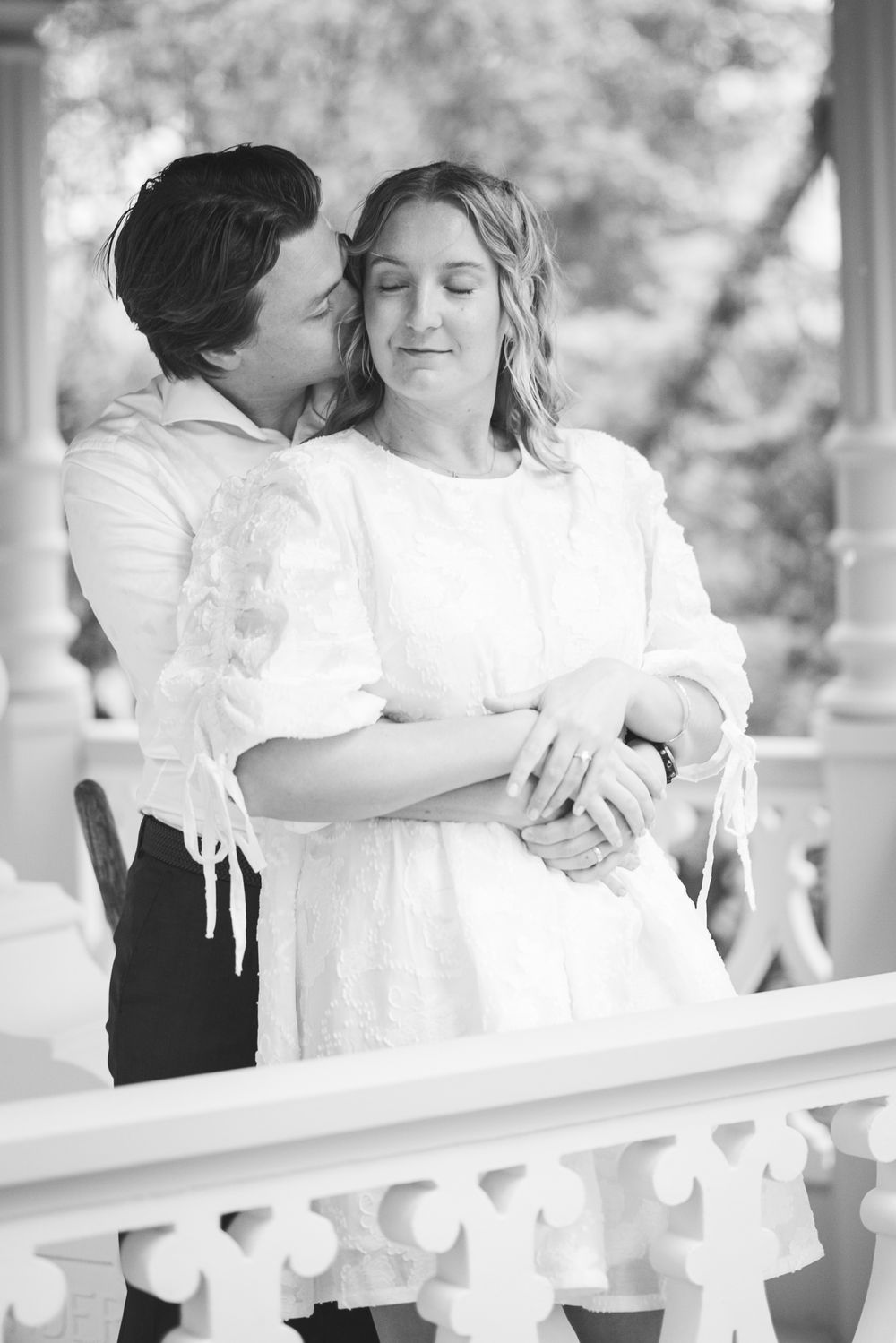 A romantic black and white wedding moment captured of a couple embracing on a white balcony railing.