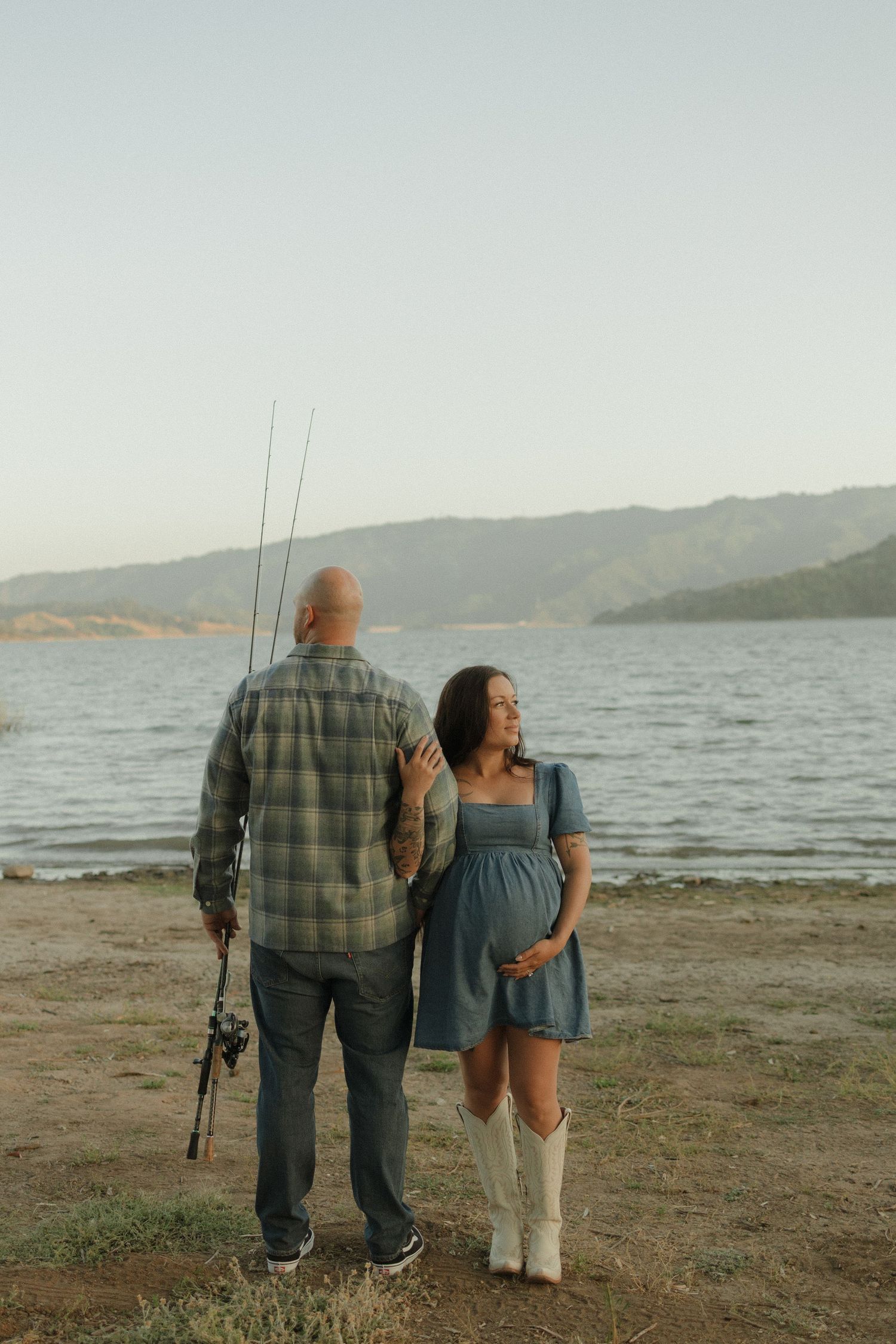 A couple holding fishing rods stands together on a beach shoreline overlooking a serene lake with mountains in the distance.