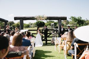 Wedding guests gather outdoors under a rustic wooden pergola for a sunny countryside celebration.