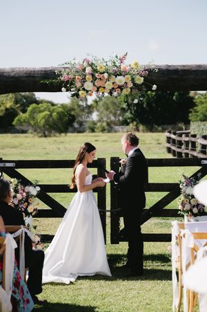 Outdoor wedding ceremony at rustic wooden gate decorated with romantic floral arrangements and white chairs.