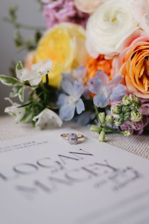 Wedding rings rest beside colorful flowers including roses and delphinium on a printed surface.